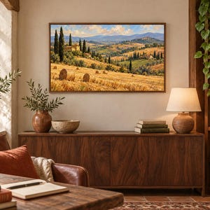 May include: A rectangular landscape painting featuring a golden wheat field with hay bales, cypress trees, and stone buildings under a blue sky with clouds. The artwork is displayed in a thin brown frame, mounted on a light-colored wall above a wooden cabinet.