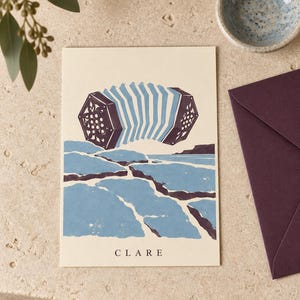 May include: A rectangular greeting card with a cream background featuring a dark purple and light blue illustration of an accordion floating above a stylized blue and purple landscape of cracked earth. The word CLARE is printed in black at the bottom.