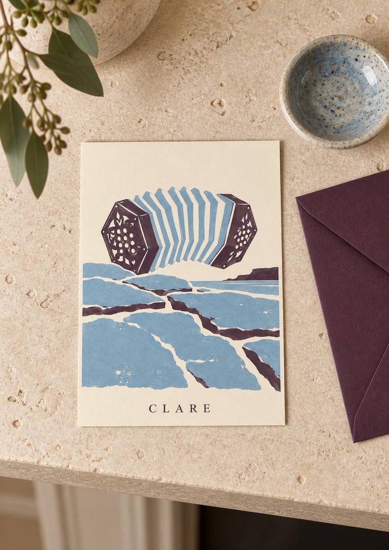 May include: A rectangular greeting card with a cream background featuring a dark purple and light blue illustration of an accordion floating above a stylized blue and purple landscape of cracked earth. The word CLARE is printed in black at the bottom.