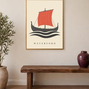 Pode incluir: Uma impress&atilde;o de arte de parede emoldurada apresentando um navio viking preto estilizado com uma vela vermelha vibrante, navegando sobre ondas pretas em um fundo creme. A palavra WATERFORD est&aacute; impressa em letras mai&uacute;sculas pretas abaixo.