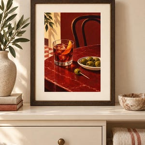 May include: A framed art print depicting a dark red table with a glass of amber liquid garnished with an orange peel, a small white bowl filled with green olives, and a single olive on a toothpick, set against a deep red wall and chair.