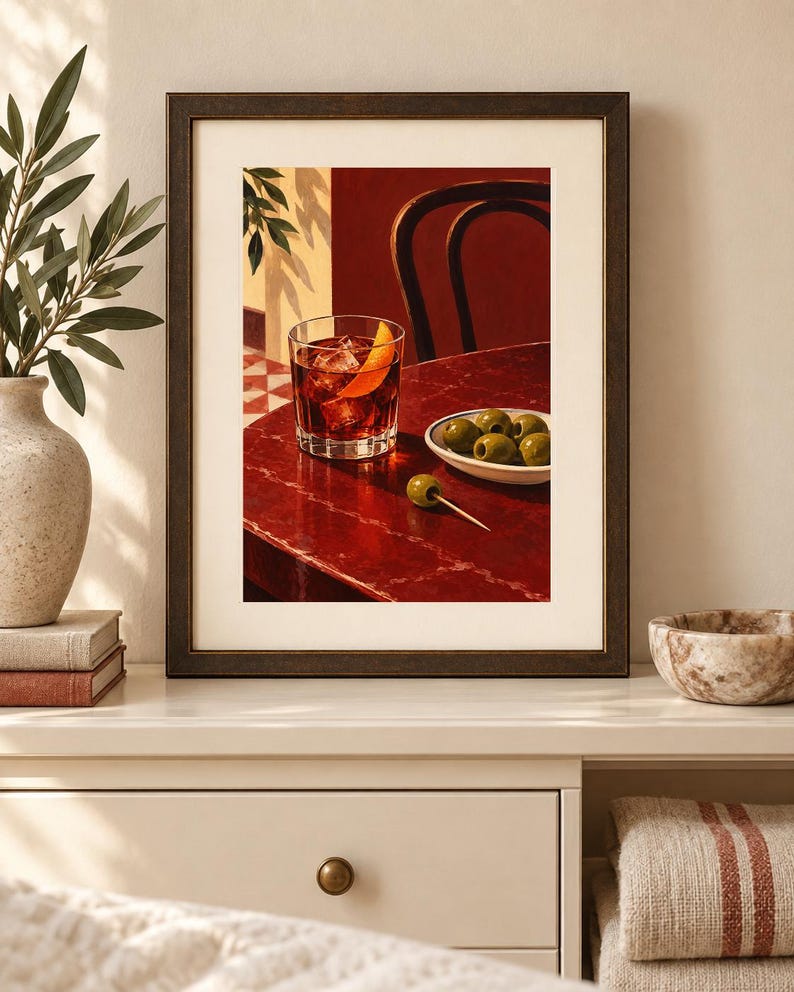 May include: A framed art print depicting a dark red table with a glass of amber liquid garnished with an orange peel, a small white bowl filled with green olives, and a single olive on a toothpick, set against a deep red wall and chair.