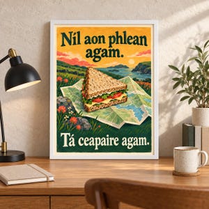 Op de afbeelding: Een ingelijste rechthoekige wandkunstprint met een driehoekige sandwich op een landkaart. De tekst luidt N&iacute;l aon phlean agam. T&aacute; ceapaire agam. De illustratie toont een kustlandschap met groene heuvels, oranje bloemen en een zonsondergang.