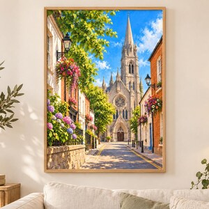 Pu&ograve; includere: Una stampa d'arte da parete incorniciata che raffigura una stretta strada europea che conduce a un'alta chiesa in pietra con una guglia. La scena presenta edifici in mattoni, cesti di fiori appesi con fiori rosa e viola, alberi verdi e un cielo blu.