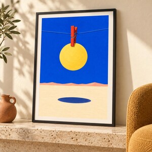 Peut inclure: Une impression d'art encadr&eacute;e repr&eacute;sentant un cercle jaune vif suspendu par une pince &agrave; linge rouge sur une ligne fine contre un ciel bleu uni. En dessous, un paysage d&eacute;sertique beige clair pr&eacute;sente une ombre ovale bleu fonc&eacute; sur le sol.