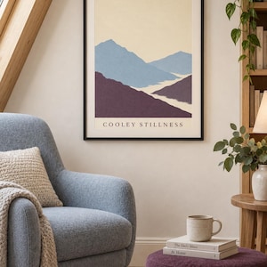 May include: A framed wall art print featuring a landscape illustration of layered mountains in shades of blue and deep purple against a pale cream background. The text "COOLEY STILLNESS" is printed in black capital letters at the bottom of the print.