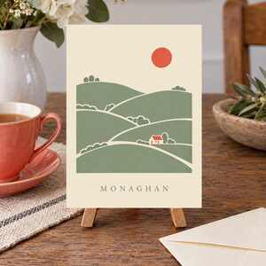 May include: A rectangular greeting card featuring a cream background with a green landscape illustration of rolling hills, a small white house with a red roof, and a solid red sun. The word MONAGHAN is printed in black capital letters at the bottom.