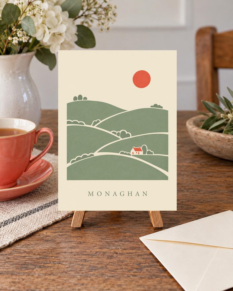 May include: A rectangular greeting card featuring a cream background with a green landscape illustration of rolling hills, a small white house with a red roof, and a solid red sun. The word MONAGHAN is printed in black capital letters at the bottom.