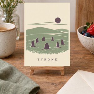 May include: A rectangular print on a small wooden easel featuring a graphic illustration of a stone circle in a green landscape under a dark purple sun. The print has a cream background and the word TYRONE printed in black capital letters at the bottom.