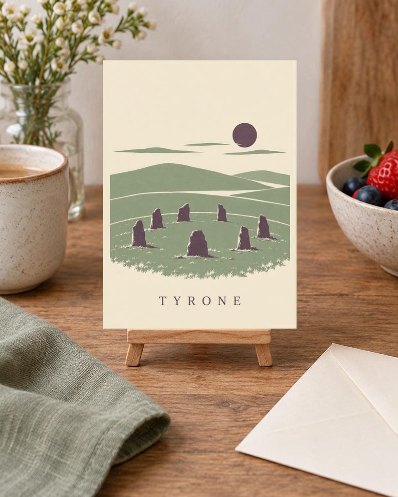 May include: A rectangular print on a small wooden easel featuring a graphic illustration of a stone circle in a green landscape under a dark purple sun. The print has a cream background and the word TYRONE printed in black capital letters at the bottom.
