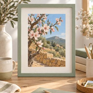 May include: A framed wall art print featuring a landscape painting of a blooming almond tree with light pink flowers in the foreground, a stone wall, and a distant mountain under a clear blue sky, enclosed in a sage green wooden frame.
