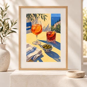 May include: A framed art print depicting a sunny outdoor scene with a tall stemmed glass of orange spritz, a short glass of red cocktail, and a small bowl of green olives on a yellow table, set against a coastal Mediterranean backdrop.