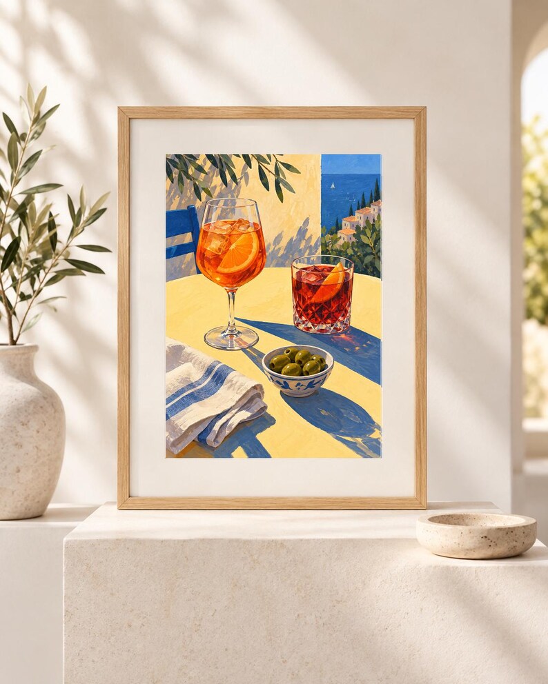 May include: A framed art print depicting a sunny outdoor scene with a tall stemmed glass of orange spritz, a short glass of red cocktail, and a small bowl of green olives on a yellow table, set against a coastal Mediterranean backdrop.