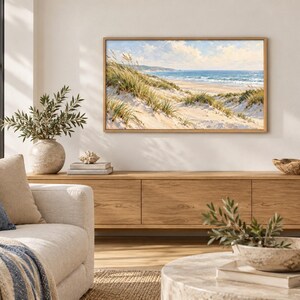 May include: A rectangular landscape painting in a light wood frame depicting a coastal scene with sand dunes, tall beach grass, and a blue ocean under a cloudy sky. The artwork is displayed on a plain wall above a long wooden media console.