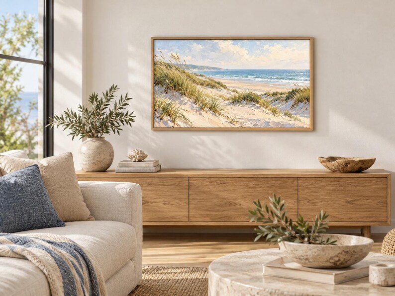 May include: A rectangular landscape painting in a light wood frame depicting a coastal scene with sand dunes, tall beach grass, and a blue ocean under a cloudy sky. The artwork is displayed on a plain wall above a long wooden media console.