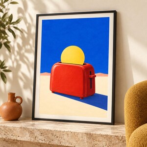 May include: A framed art print featuring a bright red toaster sitting on a light beige surface against a vibrant blue sky. A large, solid yellow circle emerges from the toaster slot, resembling a sun rising over a desert landscape.