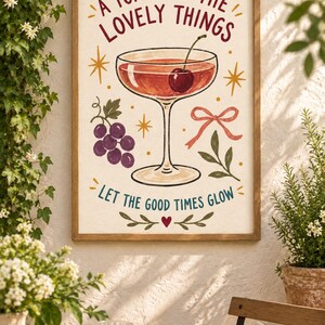 May include: A framed wall art print featuring a cocktail glass with a cherry, a cluster of purple grapes, a pink ribbon, and green leaves. The text reads A TOAST TO THE LOVELY THINGS and LET THE GOOD TIMES GLOW, accented by yellow star icons and a small red heart.