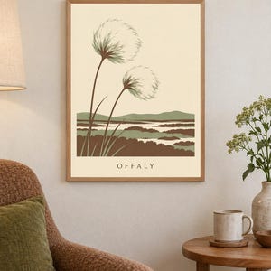 May include: A framed rectangular wall art print featuring a cream background with two stylized fluffy seed heads on long stems. Below the plants is a landscape of brown and green hills and water. The word OFFALY is printed in black at the bottom.