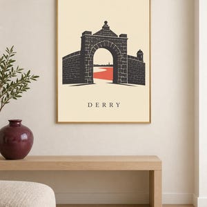May include: A framed wall art print featuring a stylized illustration of a stone archway and city walls in dark gray against a cream background. A red path leads through the archway toward a distant horizon. The word DERRY is printed in black.