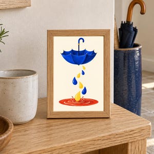 Pu&ograve; includere: Una cornice rettangolare in legno contenente la stampa di un ombrello blu sospeso in aria. Sotto l'ombrello, gocce blu e gialle cadono in una pozza rossa circolare, creando un effetto di schizzo su uno sfondo color crema chiaro.