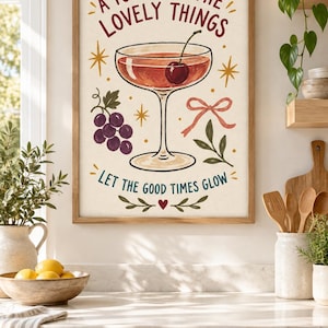 May include: A framed wall art print featuring a cocktail glass with a cherry, a cluster of purple grapes, a pink ribbon, and gold star accents. The text reads A TOAST TO THE LOVELY THINGS and LET THE GOOD TIMES GLOW, with a small red heart at the bottom.