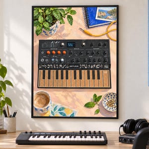 May include: A framed art print depicting an Arturia MicroFreak synthesizer with a black chassis, orange knobs, and a textured brown keyboard. The illustration includes a potted plant, a blue notebook, a coffee cup, and paper clips on a table.