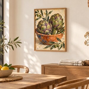 May include: A framed wall art print featuring an oil painting of two green artichokes resting in a rustic, textured terracotta-colored bowl, accented with small green olive branches and leaves against a neutral, light-colored background.