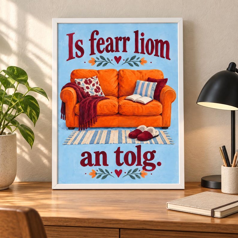 K&ouml;nnte beinhalten: Ein gerahmter Druck mit einem orangefarbenen Sofa auf blauem Hintergrund und dem Text Is fearr liom an tolg. Das Sofa hat eine kastanienbraune Decke, ein Blumenkissen, ein blau-wei&szlig; gestreiftes Kissen, ein offenes Buch und rote Hausschuhe auf einem Teppich.