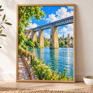 May include: A framed landscape painting depicting a stone and metal railway viaduct spanning a wide blue river. The scene includes a sunlit stone walkway with green foliage and flowers in the foreground, set against a bright blue sky with clouds.