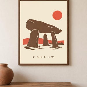 May include: A framed wall art print featuring a brown megalithic dolmen structure against a cream background. The design includes a solid red sun, red hills, and the text CARLOW printed in brown at the bottom center of the rectangular composition.