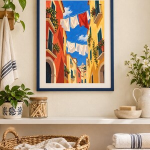 Op de afbeelding: Een ingelijste kunstdruk met een levendig straatbeeld tussen hoge rode en gele gebouwen, met wasgoed aan lijnen tegen een helderblauwe lucht. Het kunstwerk is geplaatst in een donkerblauwe lijst en vangt een zonnige mediterrane sfeer.