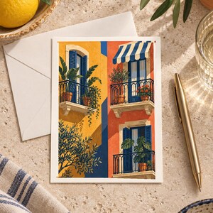 May include: A rectangular art print featuring a vibrant illustration of Mediterranean-style building facades with yellow and orange walls, blue shutters, black iron balconies, potted plants, and a blue and white striped awning over one window.