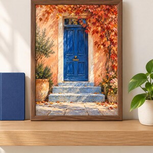 May include: A framed wall art print depicting a vibrant blue door set into a peach-coloured wall. The scene features stone steps, a potted olive tree, and climbing autumn foliage with red and orange leaves. The artwork is encased in a dark wood frame.