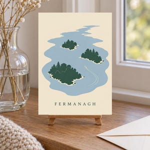 May include: A rectangular print on a small wooden easel featuring a light cream background with a blue lake illustration containing three dark green islands with trees and a small white boat leaving a wake. The word FERMANAGH is printed in black.