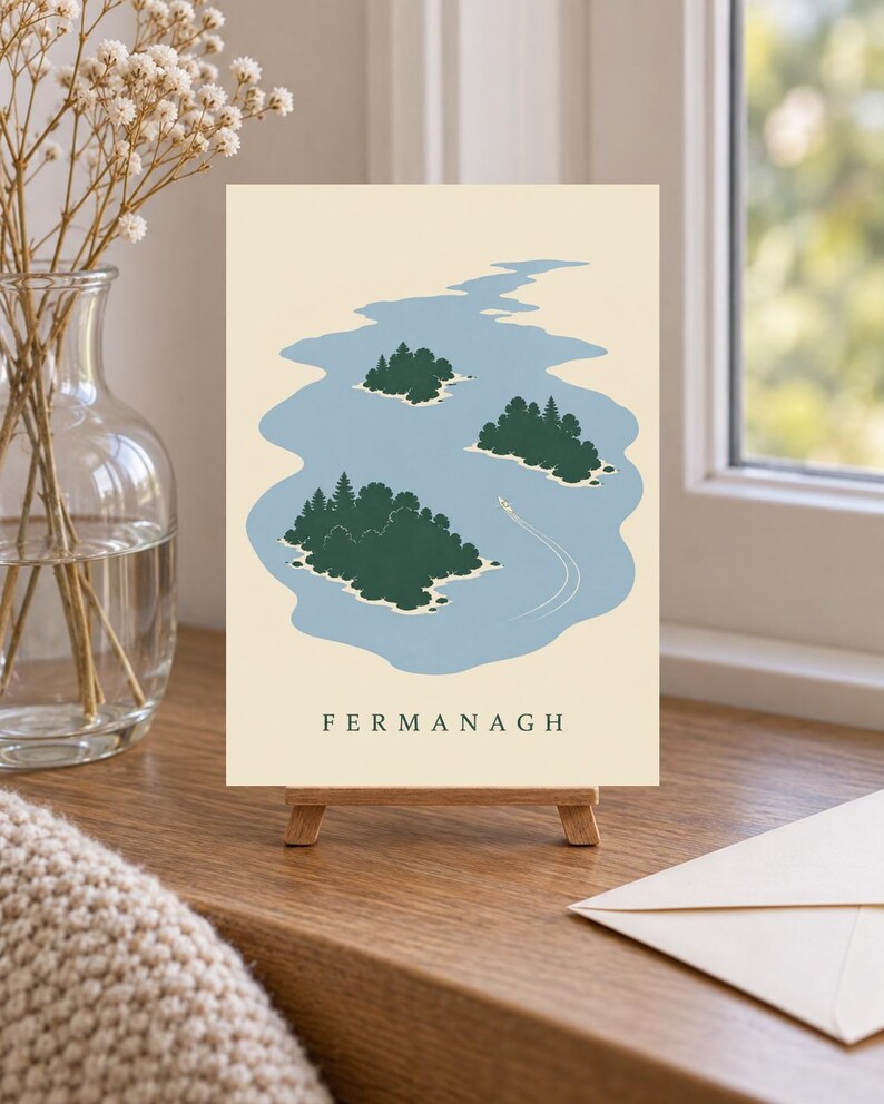 May include: A rectangular print on a small wooden easel featuring a light cream background with a blue lake illustration containing three dark green islands with trees and a small white boat leaving a wake. The word FERMANAGH is printed in black.