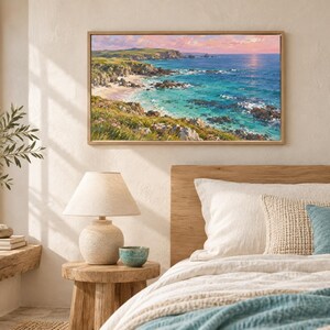May include: A rectangular landscape painting in a light wood frame depicting a coastal scene with turquoise water, rocky cliffs, and green hills under a soft pink and orange sunset sky. The artwork is displayed on a neutral wall above a bed.