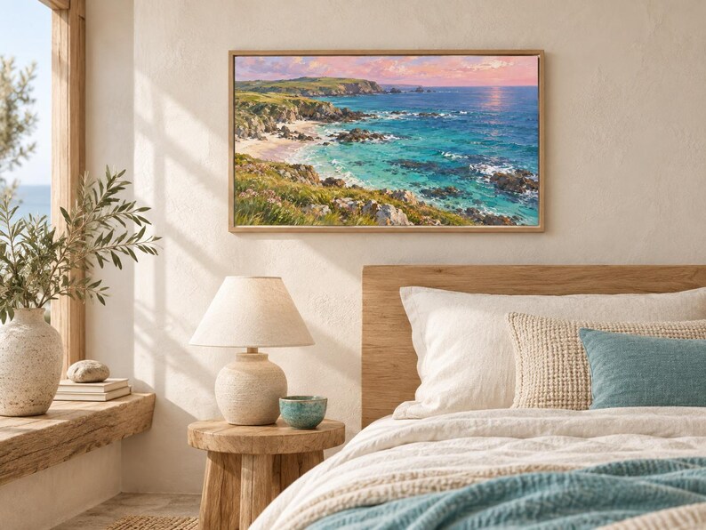May include: A rectangular landscape painting in a light wood frame depicting a coastal scene with turquoise water, rocky cliffs, and green hills under a soft pink and orange sunset sky. The artwork is displayed on a neutral wall above a bed.