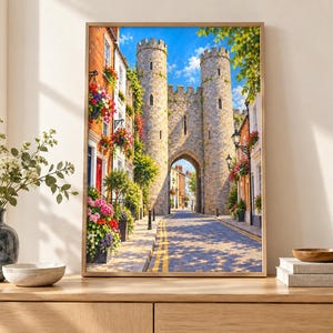 Op de afbeelding: Een ingelijste verticale wandkunstprint met een middeleeuws stenen poortgebouw met twee torens over een smalle straat. De sc&egrave;ne bevat kleurrijke bloembakken aan bakstenen gebouwen, een blauwe lucht en een geplaveide weg in het zonlicht.
