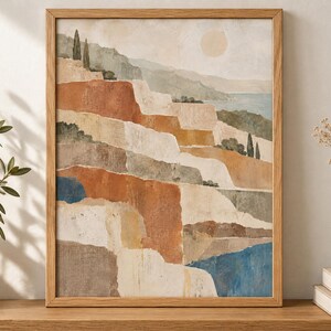 Op de afbeelding: Een ingelijste kunstdruk met een gelaagd landschap van abstracte terrasvormige kliffen in tinten terracotta, beige en saliegroen, tegen een bleke lucht met een cirkelvormige zon en een blauw wateroppervlak in de verte.