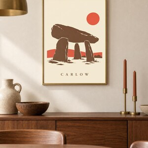 May include: A framed wall art print featuring a stylized illustration of a brown megalithic dolmen structure against a cream background. A solid red circle represents the sun above, with red hills below. The text CARLOW is printed in black.