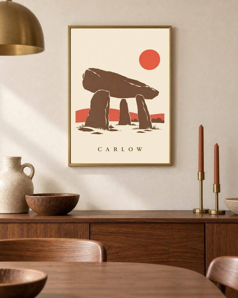 May include: A framed wall art print featuring a stylized illustration of a brown megalithic dolmen structure against a cream background. A solid red circle represents the sun above, with red hills below. The text CARLOW is printed in black.