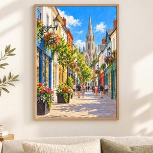 Op de afbeelding: Een ingelijste wandkunstprint met een zonnig Europees straatbeeld met kleurrijke winkelpuien, hangende bloemenmanden en een hoge stenen kerktoren op de achtergrond. Mensen lopen over het kasseienpad onder een helderblauwe lucht.