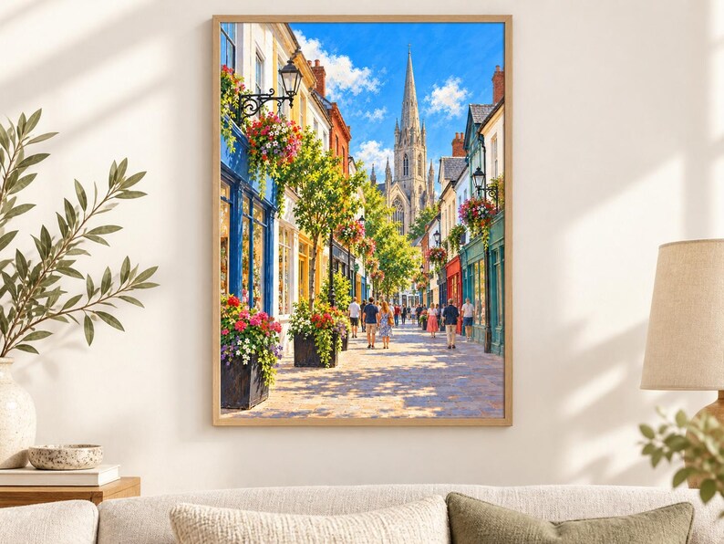 Op de afbeelding: Een ingelijste wandkunstprint met een zonnig Europees straatbeeld met kleurrijke winkelpuien, hangende bloemenmanden en een hoge stenen kerktoren op de achtergrond. Mensen lopen over het kasseienpad onder een helderblauwe lucht.