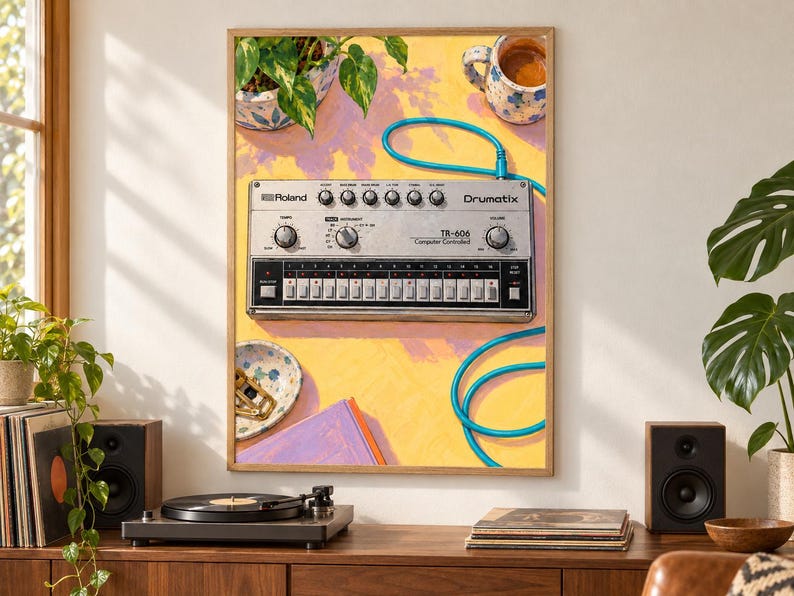 Pu&ograve; includere: Una stampa artistica incorniciata che raffigura una drum machine Roland TR-606 Drumatix argentata su una superficie gialla. L'illustrazione include un cavo audio blu, una pianta in vaso, una tazza in ceramica, una ciotola e un libro viola.
