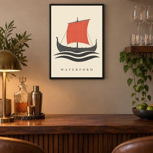 Pode incluir: Uma impress&atilde;o art&iacute;stica emoldurada com um navio viking preto estilizado com uma vela vermelha vibrante sobre um fundo creme. A ilustra&ccedil;&atilde;o mostra o navio sobre linhas escuras e onduladas, com a palavra WATERFORD impressa abaixo.