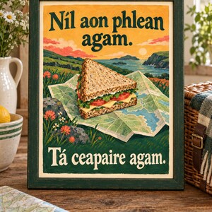 Op de afbeelding: Een ingelijste rechthoekige print met een driehoekige sandwich op een kaart. Het kunstwerk toont een kustlandschap met oranje bloemen en de tekst N&iacute;l aon phlean agam en T&aacute; ceapaire agam in een donkergroen schreeflettertype.