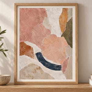 May include: A framed abstract wall art print featuring a collage of organic shapes in muted tones of dusty pink, terracotta, cream, olive green, and a dark navy blue curved accent, all set within a light-toned wooden frame with a white mat border.