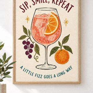 Op de afbeelding: Een ingelijste kunstprint met een cocktail in een glas op voet met ijsblokjes en een schijfje sinaasappel. De tekst luidt SIP, SMILE, REPEAT bovenaan en A LITTLE FIZZ GOES A LONG WAY onderaan. Illustraties van paarse druiven en een sinaasappel.