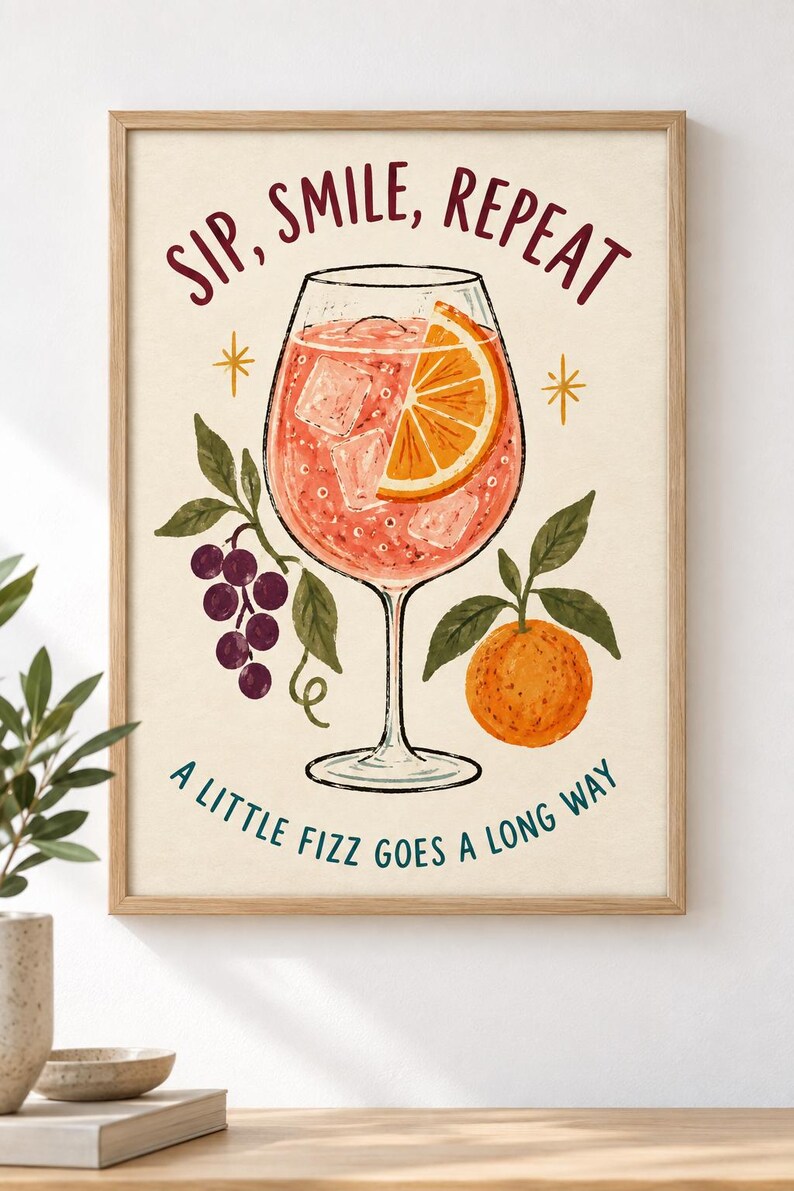 Op de afbeelding: Een ingelijste kunstprint met een cocktail in een glas op voet met ijsblokjes en een schijfje sinaasappel. De tekst luidt SIP, SMILE, REPEAT bovenaan en A LITTLE FIZZ GOES A LONG WAY onderaan. Illustraties van paarse druiven en een sinaasappel.