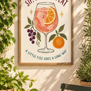 Op de afbeelding: Een ingelijste kunstprint met een glas roze cocktail met ijs en een schijfje sinaasappel. De tekst luidt bovenaan SIP, SMILE, REPEAT en onderaan A LITTLE FIZZ GOES A LONG WAY. Illustraties van paarse druiven en een sinaasappel zijn inbegrepen.